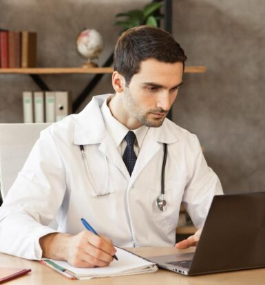 Doctor in a white coat working on a laptop and taking notes to manage healthcare administrative tasks efficiently.