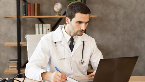 Doctor in a white coat working on a laptop and taking notes to manage healthcare administrative tasks efficiently.