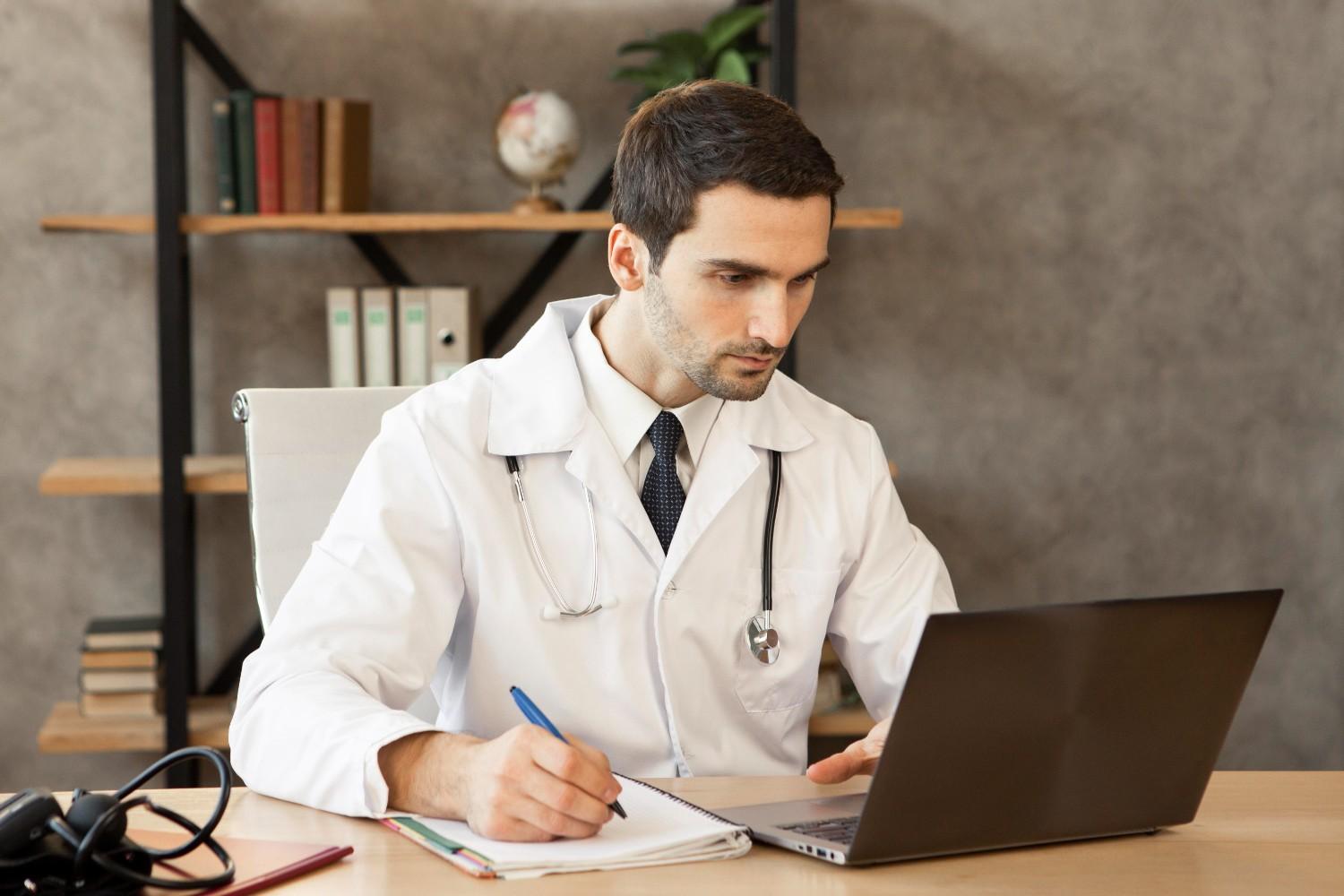 Doctor in a white coat working on a laptop and taking notes to manage healthcare administrative tasks efficiently.