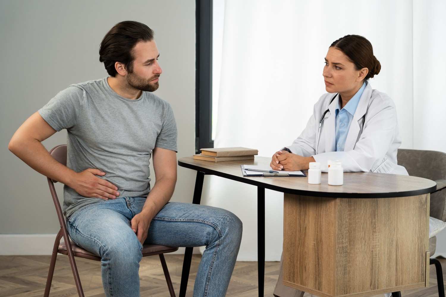 A concerned male patient holding his abdomen, sitting across from a doctor who is attentively listening, symbolizing a consultation about Xifaxan prior authorization for IBS-D or hepatic encephalopathy.