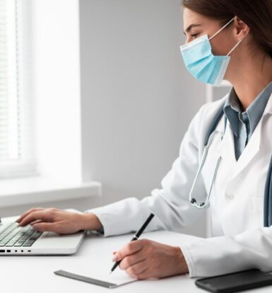 A female healthcare provider wearing a mask, working on a laptop while taking notes in a bright, professional office setting.