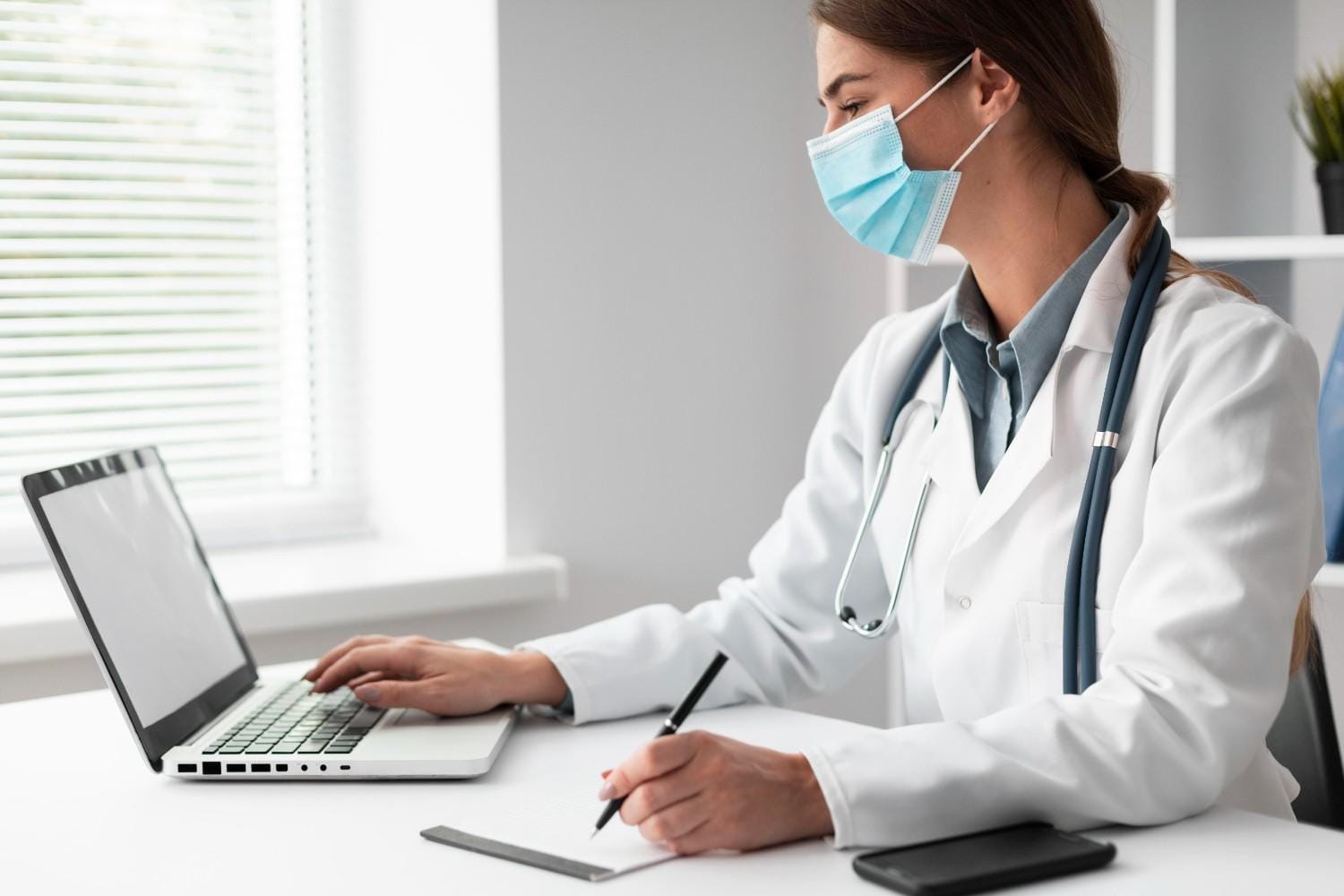 A female healthcare provider wearing a mask, working on a laptop while taking notes in a bright, professional office setting.