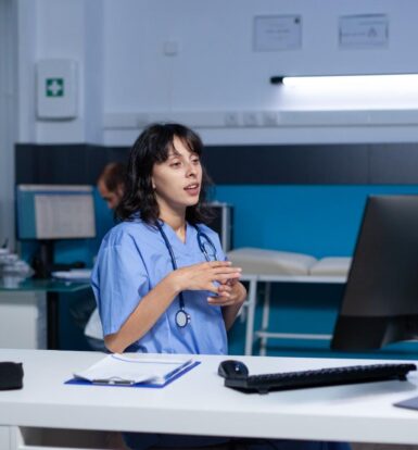 Healthcare administrator in scrubs discussing virtual assistant solutions on a video call, with a computer and documents on the desk.