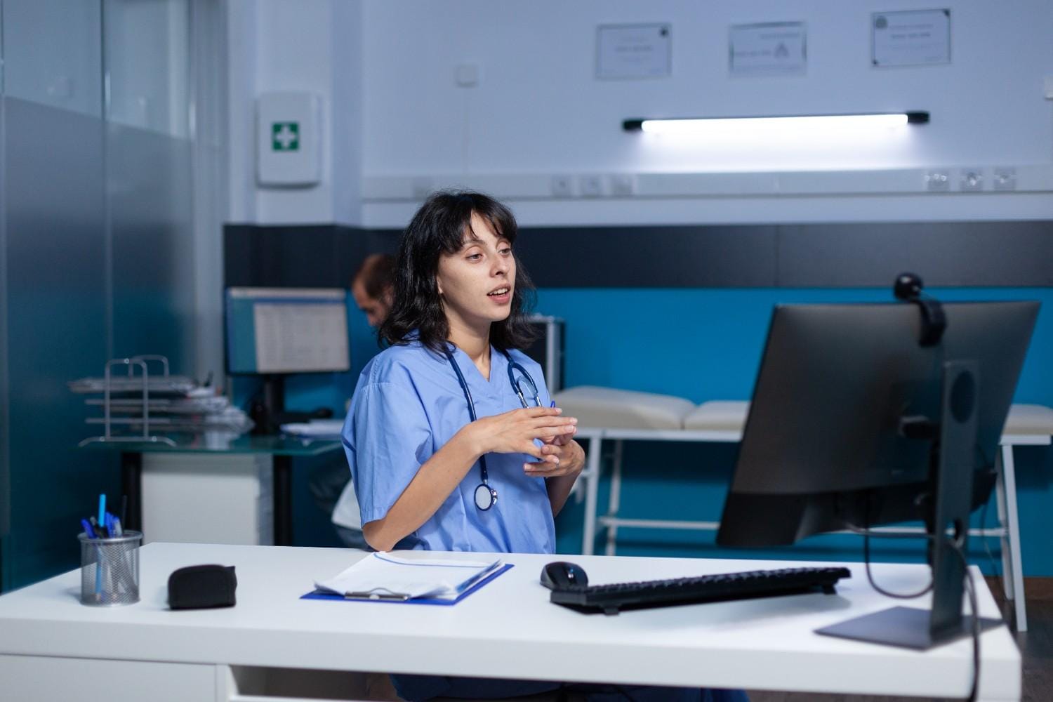 Healthcare administrator in scrubs discussing virtual assistant solutions on a video call, with a computer and documents on the desk.