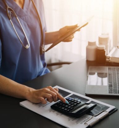 Healthcare professional in blue scrubs using a tablet and calculator at a desk with a laptop, symbolizing LTC billing and data management.