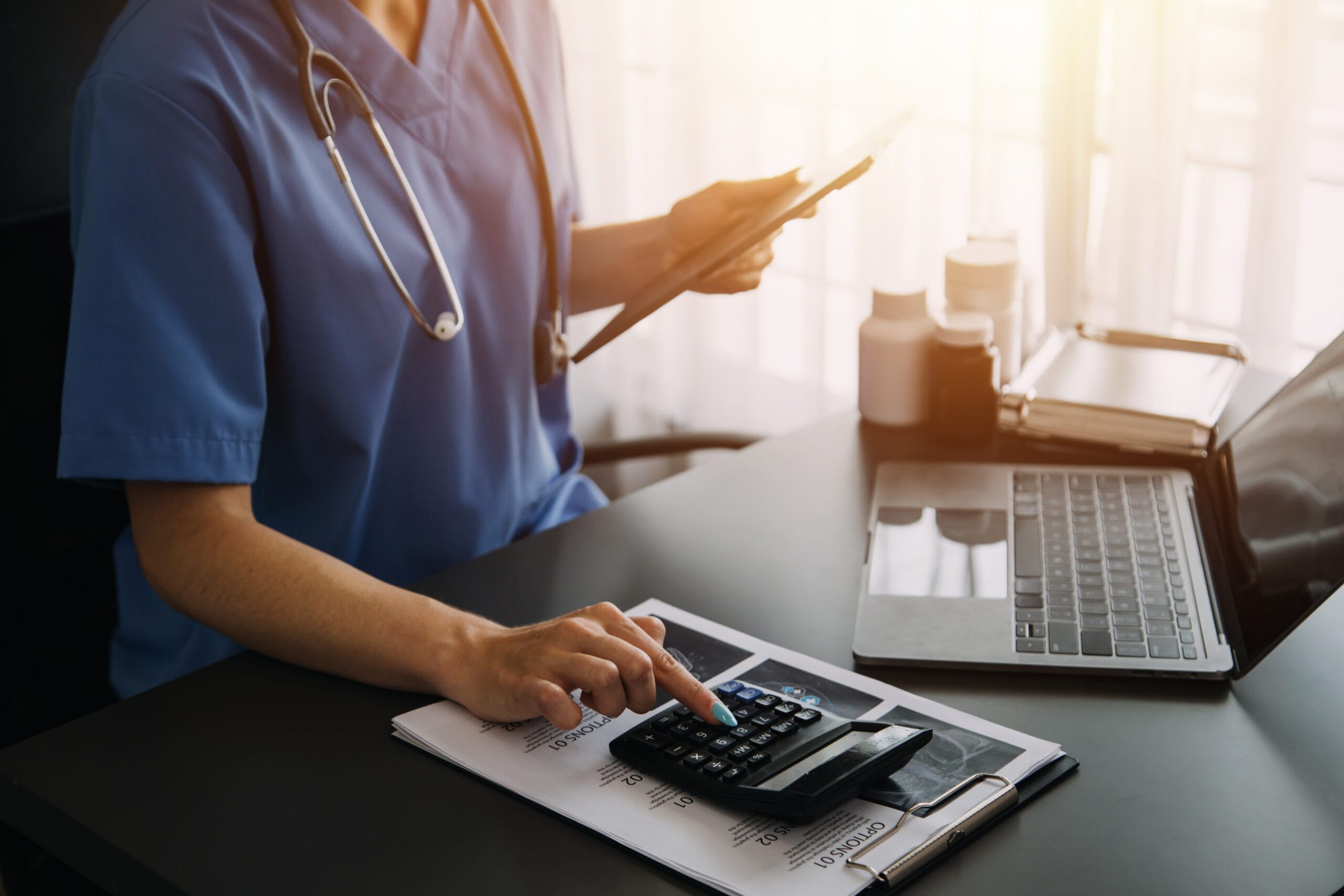 Healthcare professional in blue scrubs using a tablet and calculator at a desk with a laptop, symbolizing LTC billing and data management.