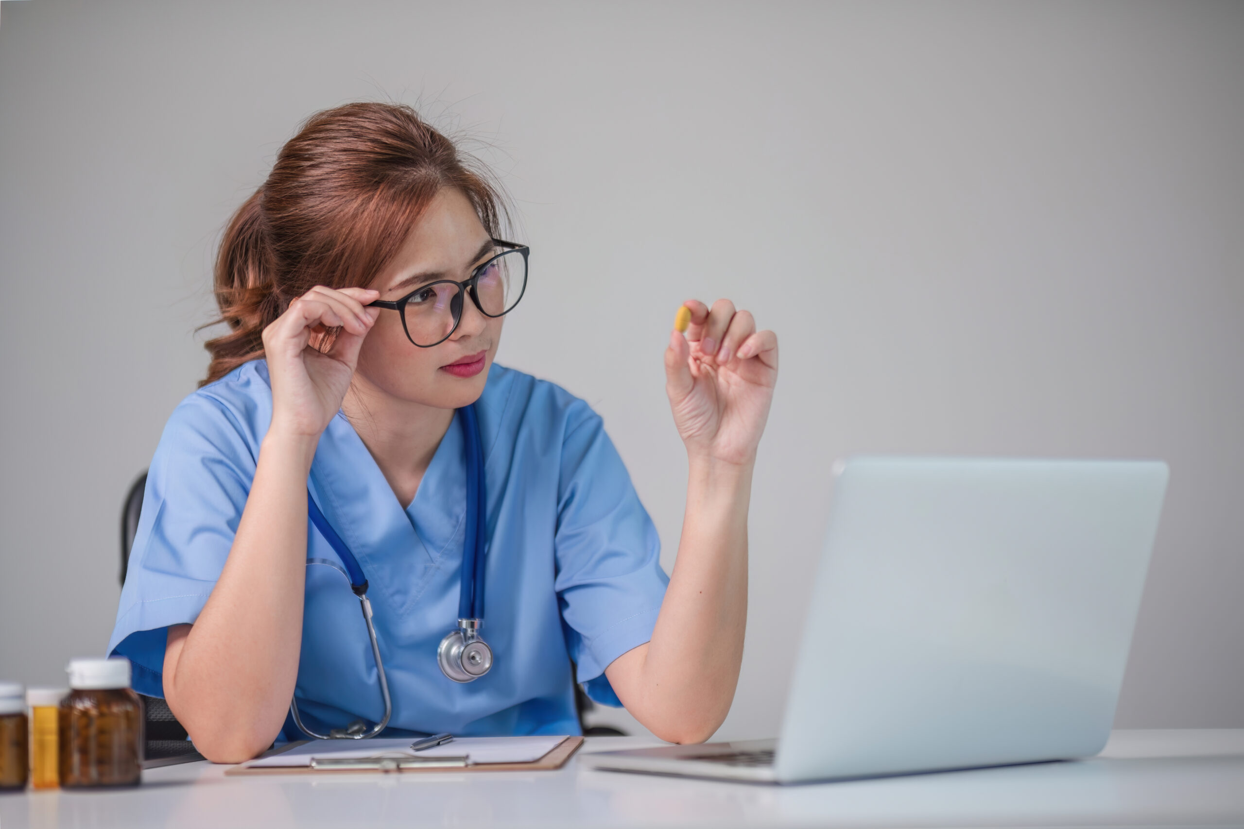 A nurse in blue scrubs sitting at a desk, examining a pill closely while working on a laptop, with bottles of medication nearby.