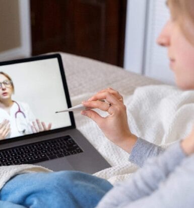 A patient holding a thermometer while consulting a virtual healthcare assistant on a laptop for medical advice.