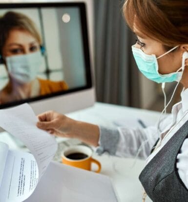 A healthcare professional wearing a mask reviewing documents during a virtual consultation with a medical assistant on a computer screen.