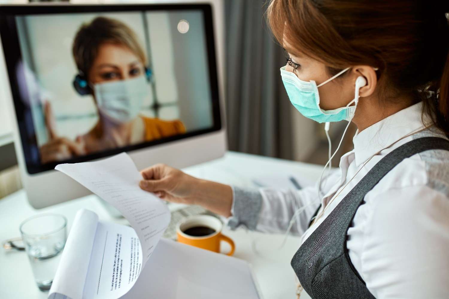 A healthcare professional wearing a mask reviewing documents during a virtual consultation with a medical assistant on a computer screen.