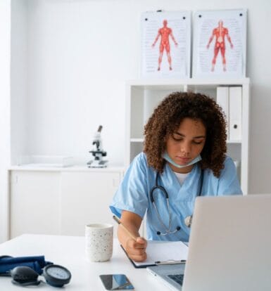 A healthcare professional using a laptop and writing notes while managing medical documentation, representing virtual medical assistants in medical scribing.