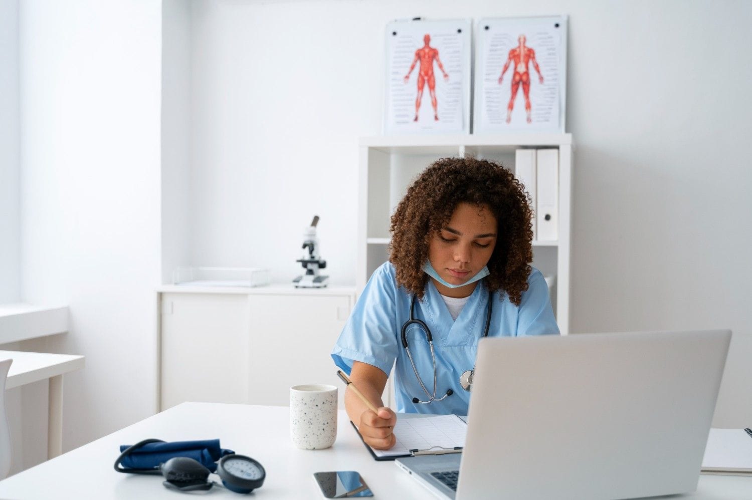 A healthcare professional using a laptop and writing notes while managing medical documentation, representing virtual medical assistants in medical scribing.