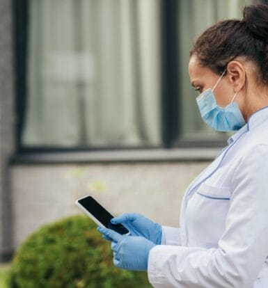 A healthcare professional in a mask and gloves reviewing patient data on a tablet outdoors.