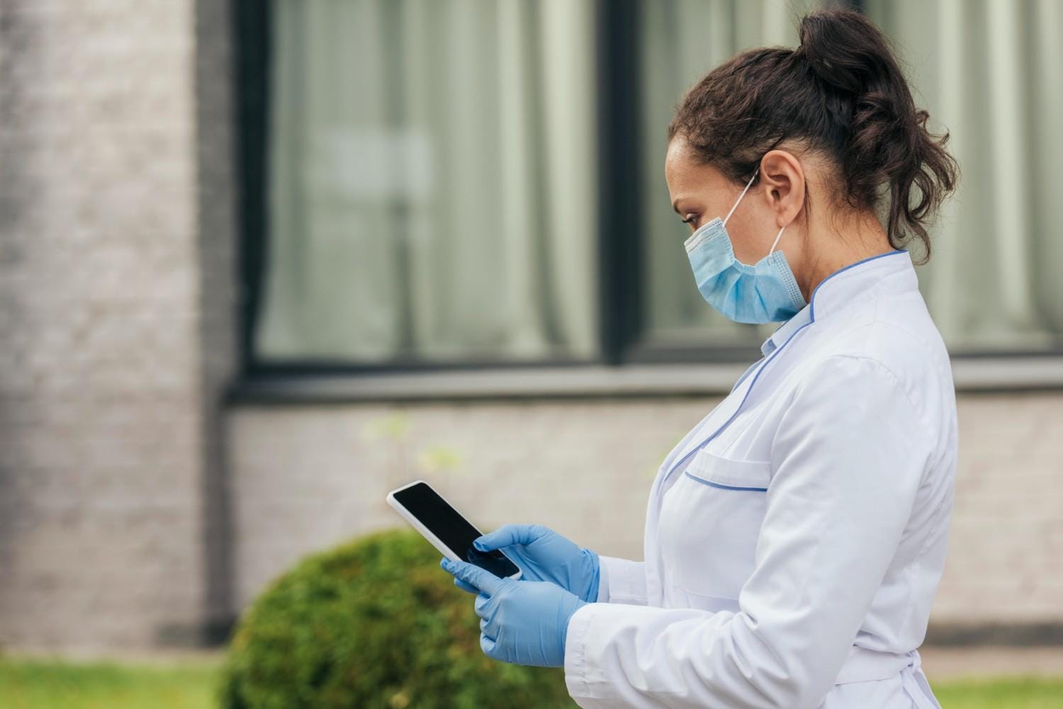 A healthcare professional in a mask and gloves reviewing patient data on a tablet outdoors.