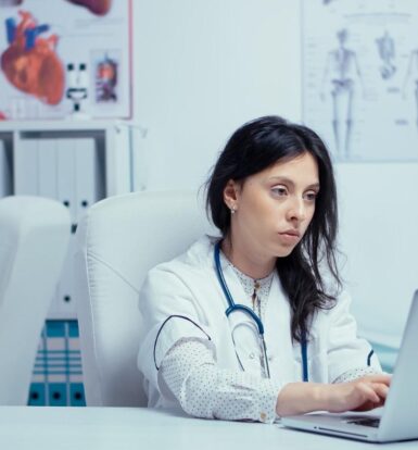 A medical professional reviewing patient data on a laptop while another staff member works in the background, showcasing effective revenue cycle management in a healthcare setting. Title: