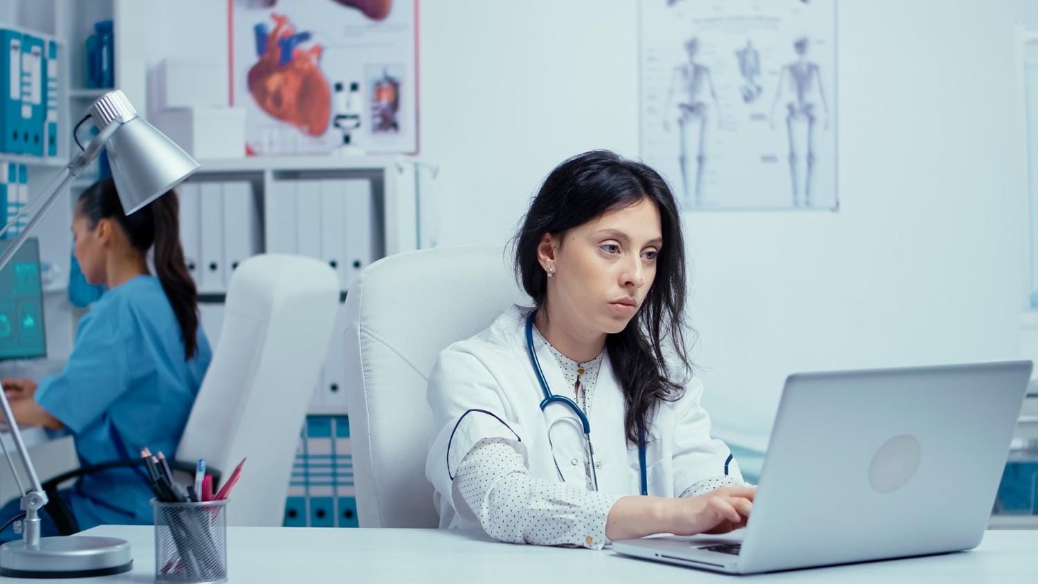 A medical professional reviewing patient data on a laptop while another staff member works in the background, showcasing effective revenue cycle management in a healthcare setting. Title: