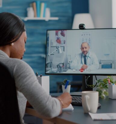 A woman working remotely as a virtual medical assistant, taking notes during a telemedicine consultation with a doctor on a computer screen.