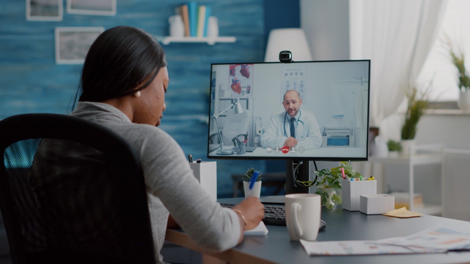 A woman working remotely as a virtual medical assistant, taking notes during a telemedicine consultation with a doctor on a computer screen.