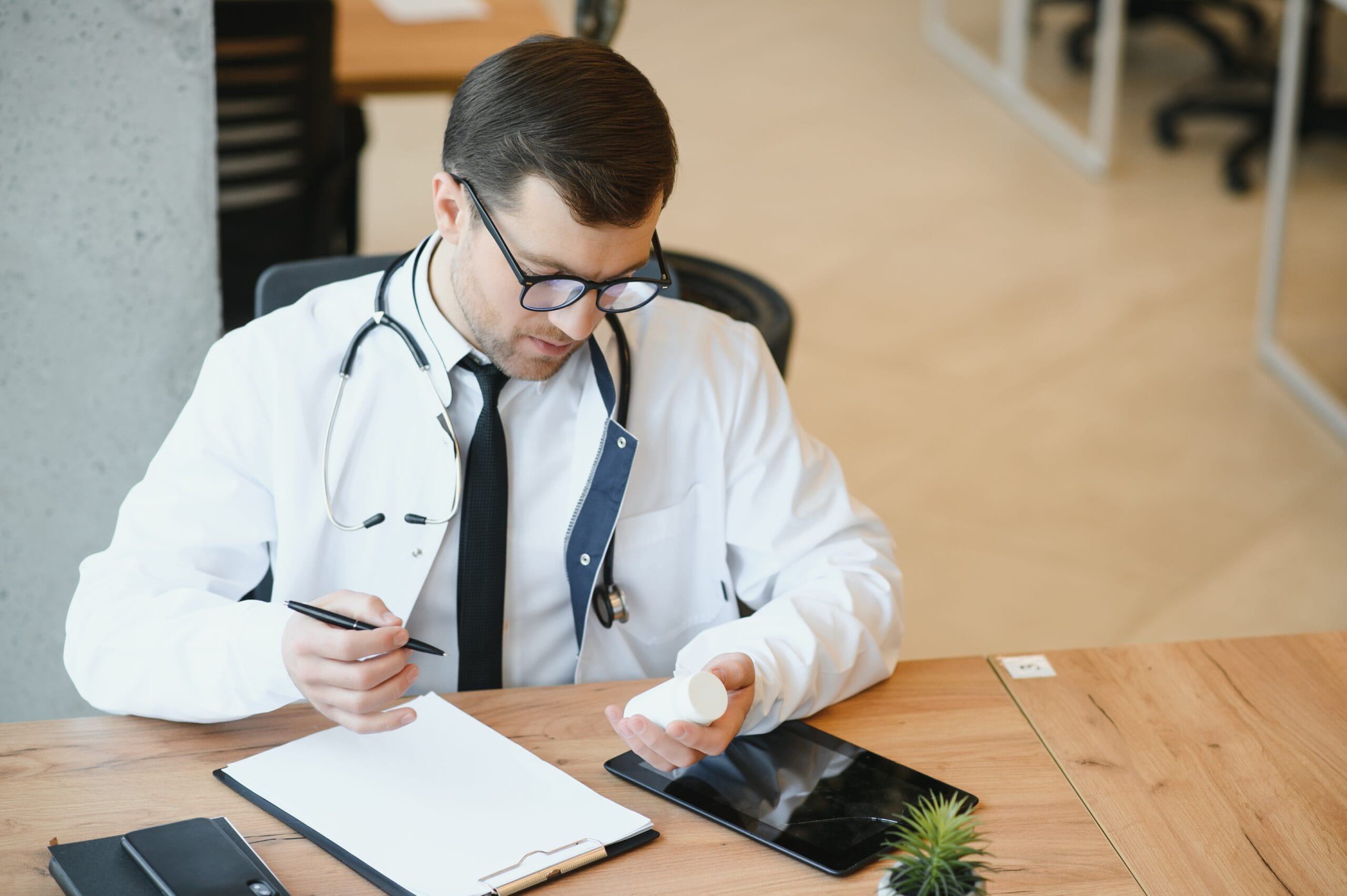 A doctor reviewing medication details while taking notes on a clipboard, with a tablet and phone on the desk.