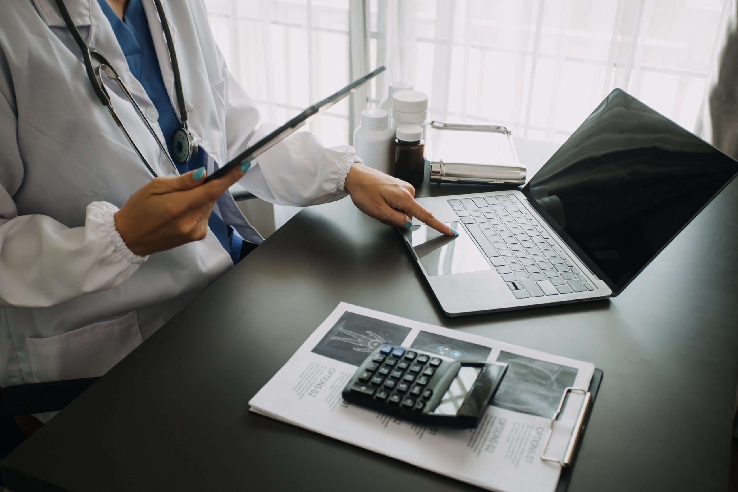 A healthcare provider using a tablet and laptop to manage EHR documentation, alongside medical tools and records on the desk.