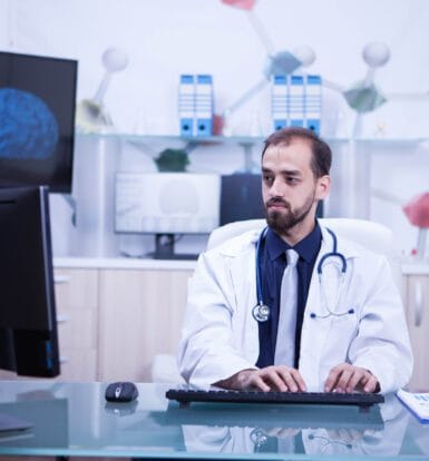 A doctor in a white coat sits at a desk using a computer, with a brain image and DNA strand displayed on the screen in the background, representing telemedicine and specialist consultations.