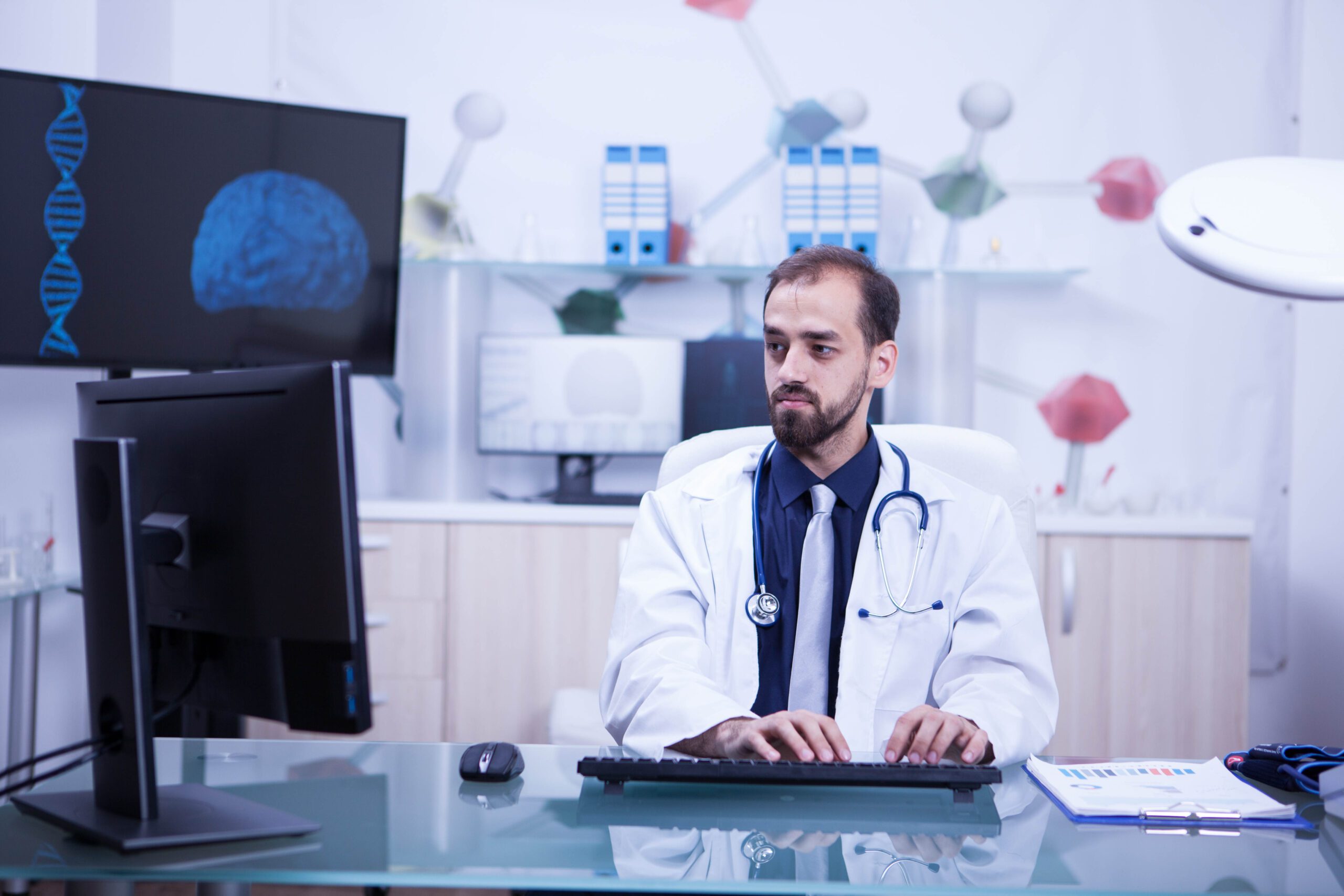 A doctor in a white coat sits at a desk using a computer, with a brain image and DNA strand displayed on the screen in the background, representing telemedicine and specialist consultations.