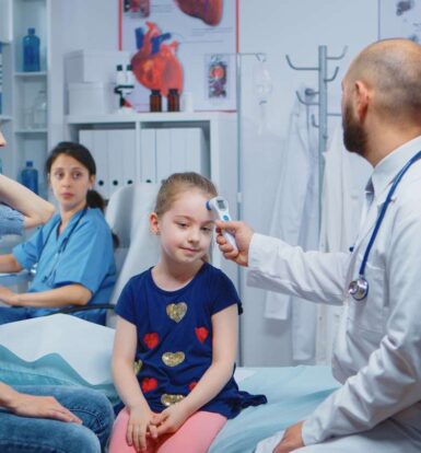 Doctor taking a young patient’s temperature in a clinical setting, with a concerned parent and nurse observing in the background.