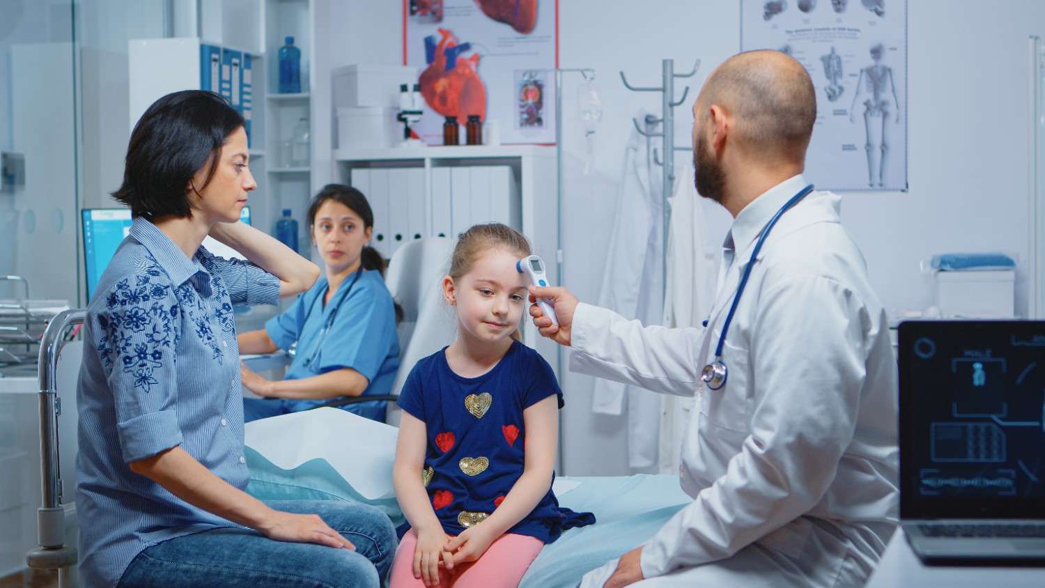 Doctor taking a young patient’s temperature in a clinical setting, with a concerned parent and nurse observing in the background.