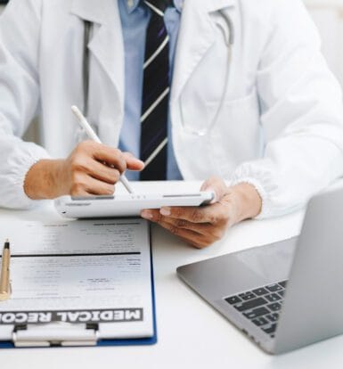 A medical scribe documenting patient notes on a computer while a doctor consults with a patient.