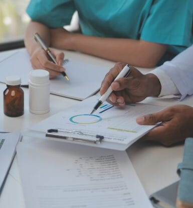 A physician interacting with a patient while a medical scribe takes notes on a laptop.