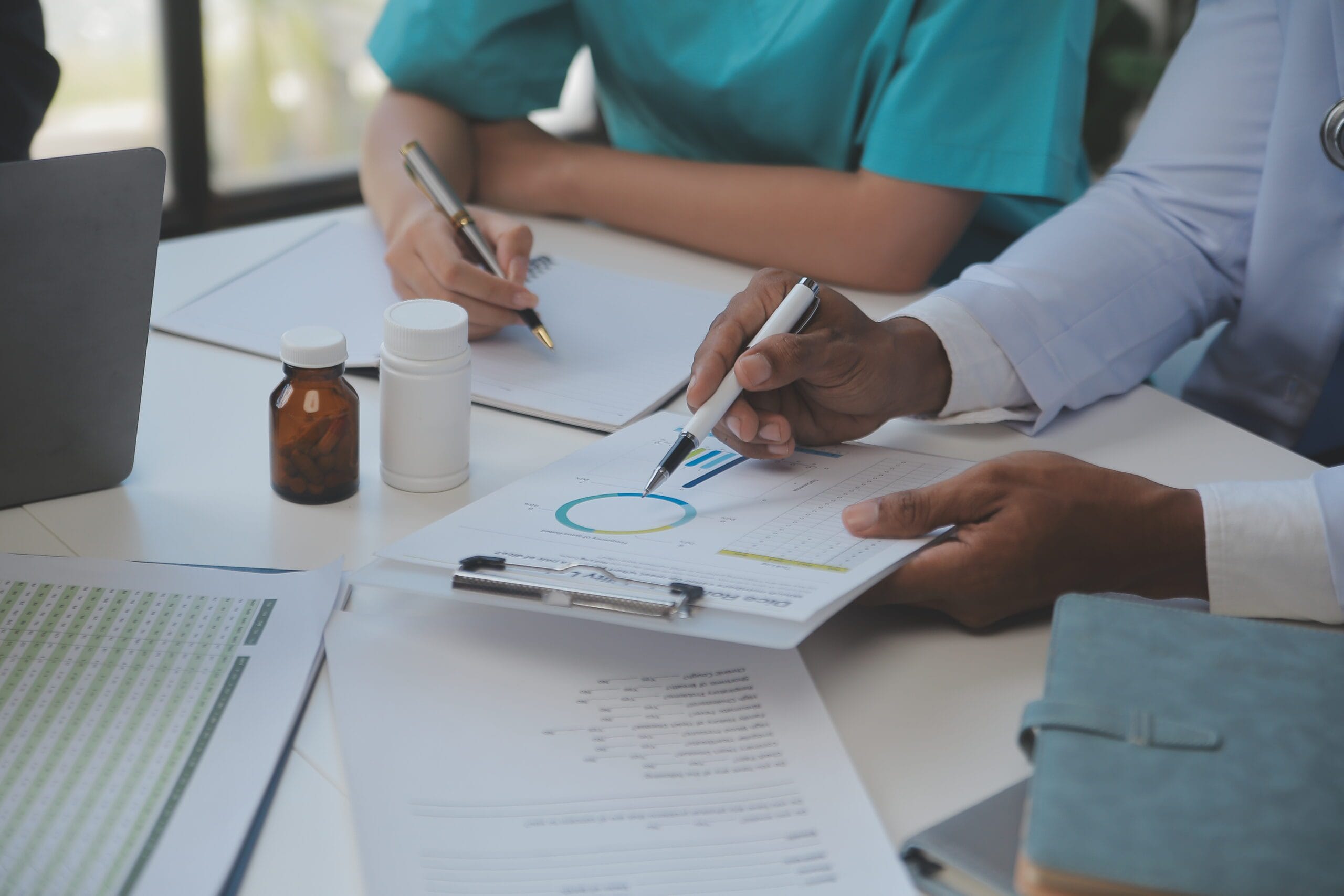 A physician interacting with a patient while a medical scribe takes notes on a laptop.