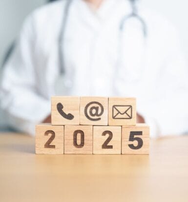 Wooden blocks with communication icons and '2025' in front of a doctor, symbolizing the future of virtual medical outsourcing services.