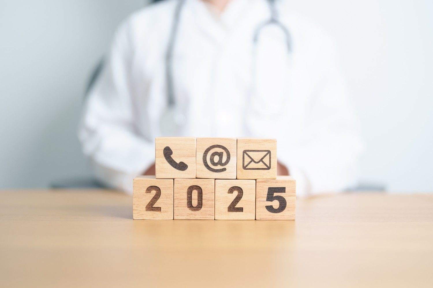 Wooden blocks with communication icons and '2025' in front of a doctor, symbolizing the future of virtual medical outsourcing services.