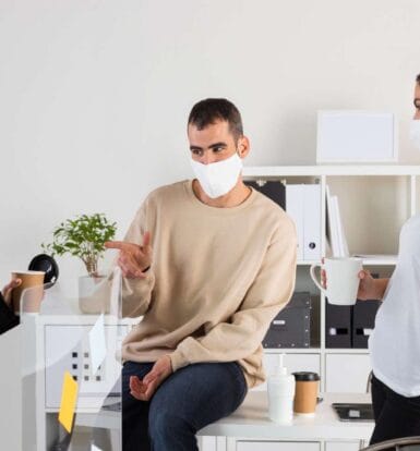 Three colleagues wearing masks discussing in an office environment, holding coffee mugs, with hand sanitizer on the table.