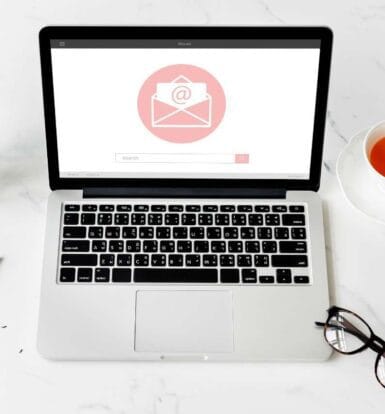 Laptop on a white desk with an email icon on the screen, surrounded by a cup of tea, glasses, flowers, and documents.