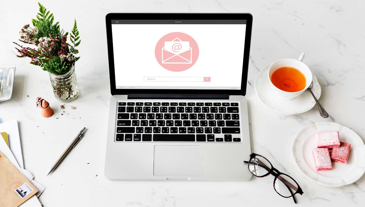 Laptop on a white desk with an email icon on the screen, surrounded by a cup of tea, glasses, flowers, and documents.