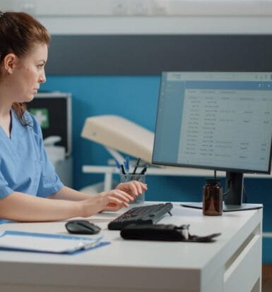 Healthcare professional in scrubs working on a computer for insurance verification and medical data entry.