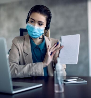 A medical outsourcing virtual assistant wearing a headset and a face mask, holding a notebook while working on a laptop remotely.