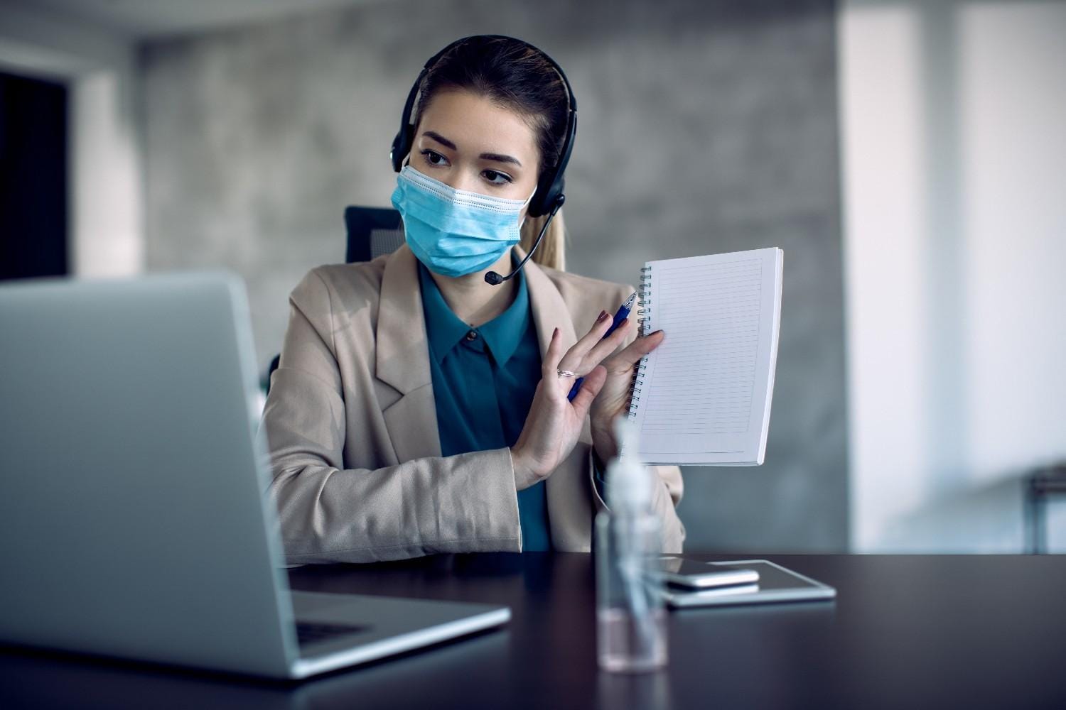 A medical outsourcing virtual assistant wearing a headset and a face mask, holding a notebook while working on a laptop remotely.