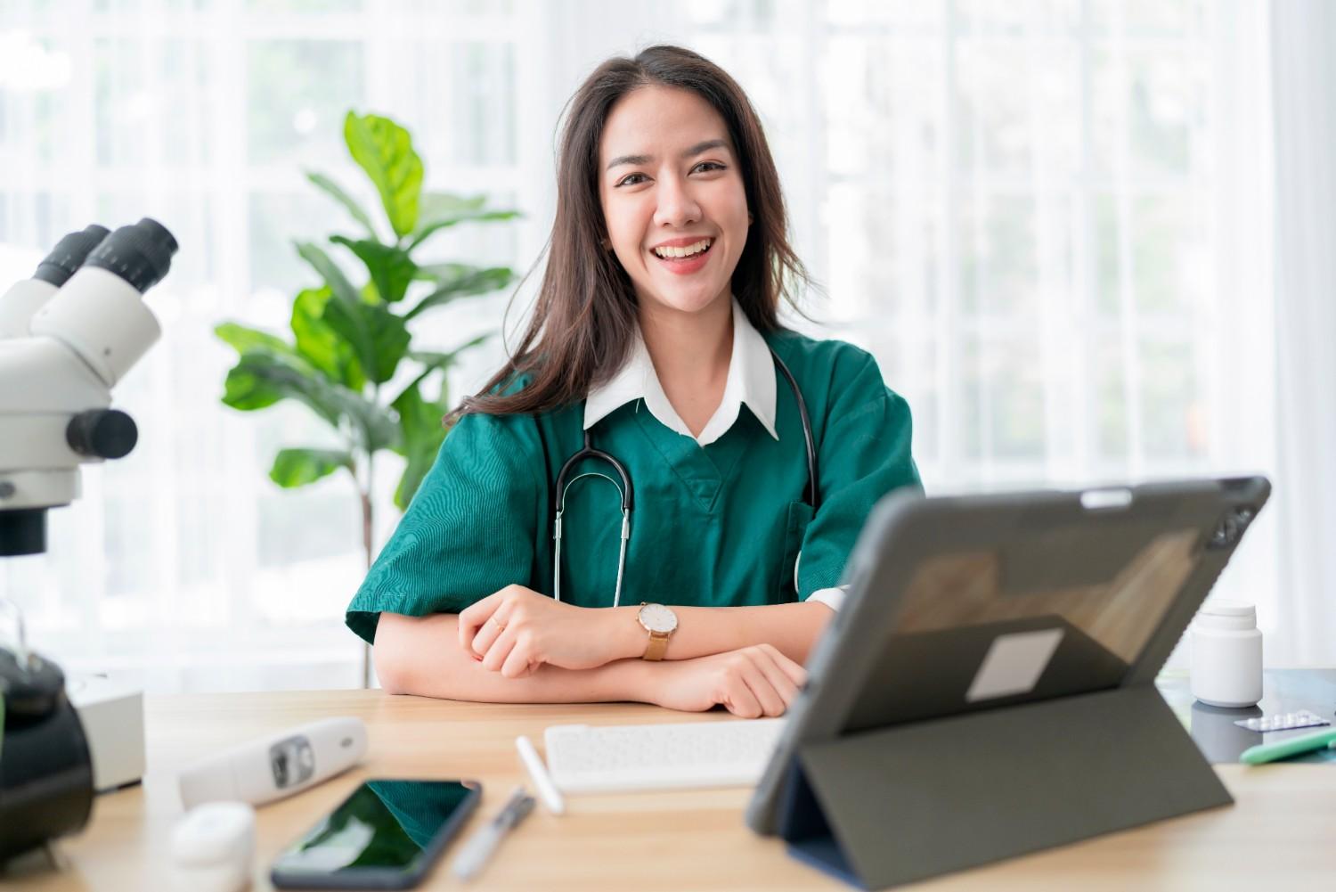 Smiling doctor in a green medical uniform using a tablet in a bright, modern healthcare setting.