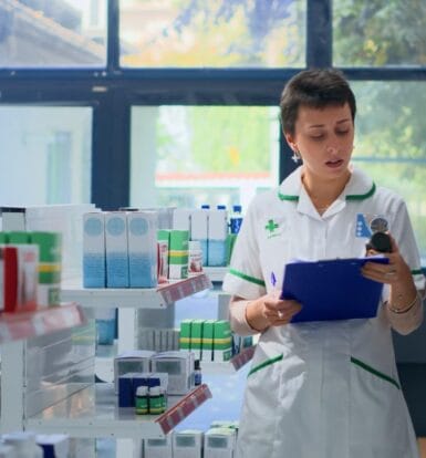 A pharmacy professional in a white uniform explaining medication options to an elderly customer, standing in a pharmacy aisle