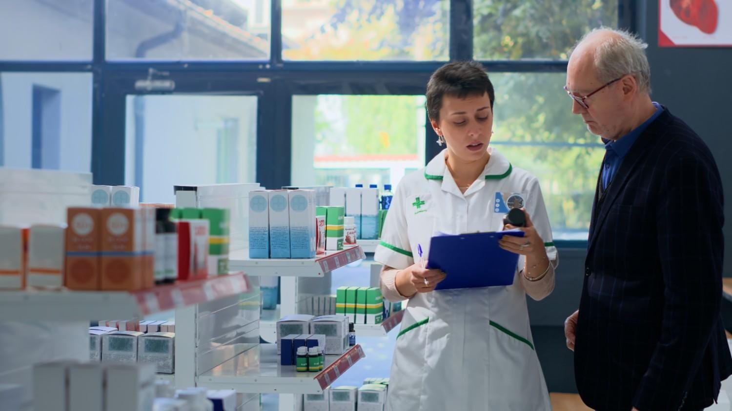 A pharmacy professional in a white uniform explaining medication options to an elderly customer, standing in a pharmacy aisle