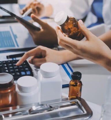 Close-up of healthcare professionals handling pharmacy billing tasks, with one person holding a medication bottle and another working on a calculator. The image includes medical forms, pill bottles, and a tablet, illustrating the complexity of LTC pharmacy billing processes.