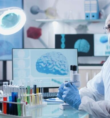 Medical researcher wearing protective gear in a laboratory with brain analysis on a screen in the background.