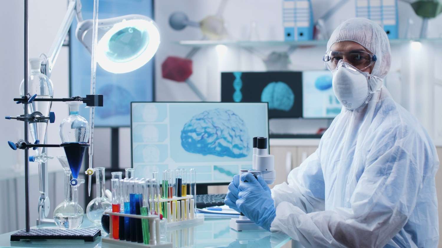 Medical researcher wearing protective gear in a laboratory with brain analysis on a screen in the background.