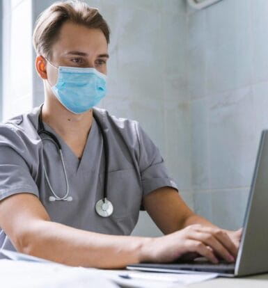 Healthcare professional wearing a mask and scrubs, working on a laptop to manage virtual medical assistant tasks remotely.