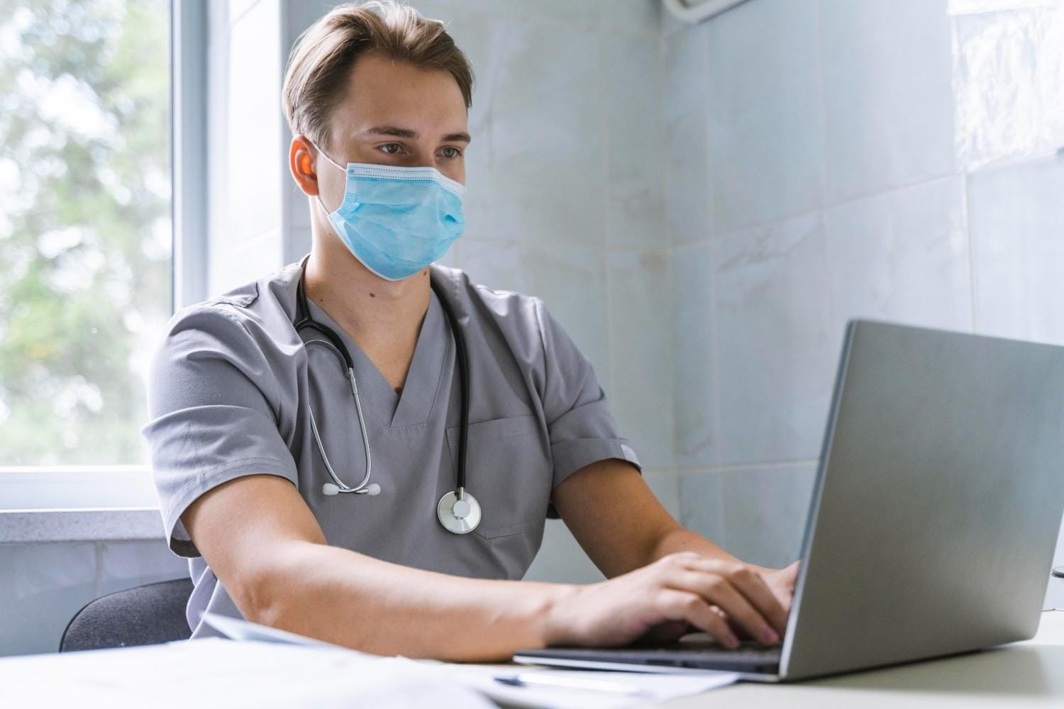 Healthcare professional wearing a mask and scrubs, working on a laptop to manage virtual medical assistant tasks remotely.