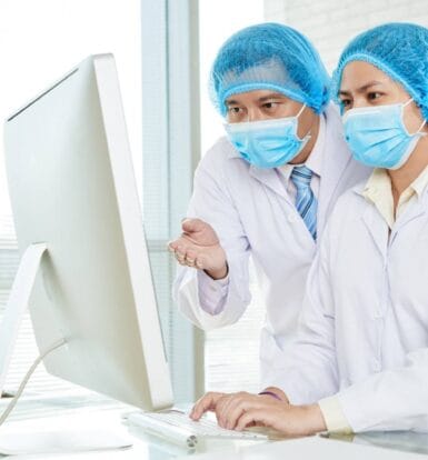 Two healthcare professionals in protective caps and face masks working together on a computer in a medical office.