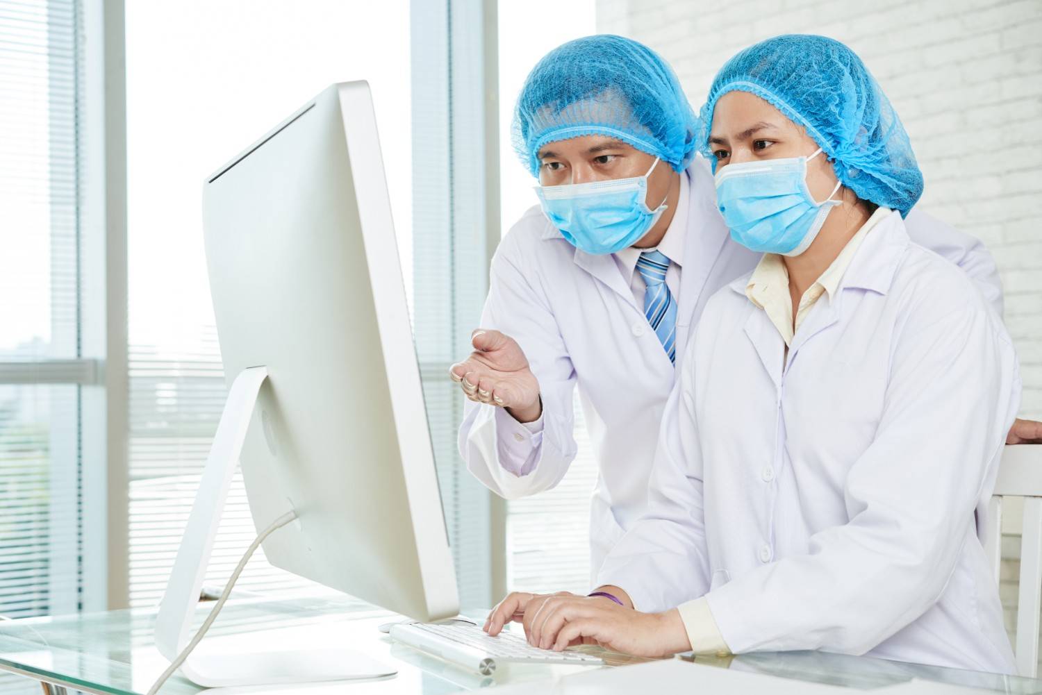 Two healthcare professionals in protective caps and face masks working together on a computer in a medical office.