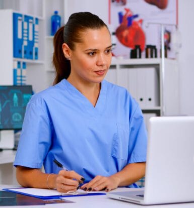 A healthcare professional in blue scrubs sitting at a desk, reviewing information on a laptop. The background shows medical equipment and anatomical charts.